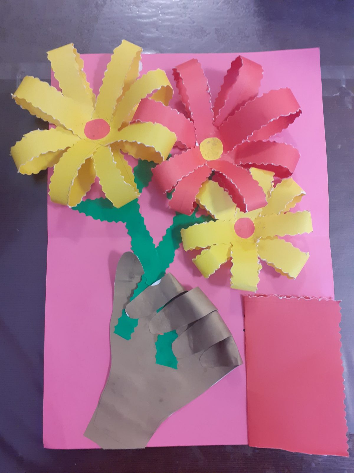 na rozowy tle ręka trzymająca 3 kwiaty - platki zewnętrznych kwiatow maja kolor żółty z różowym środkiem i czerwony na środku, z prawej na dole, kartka z ukrytym napisem dla mamy